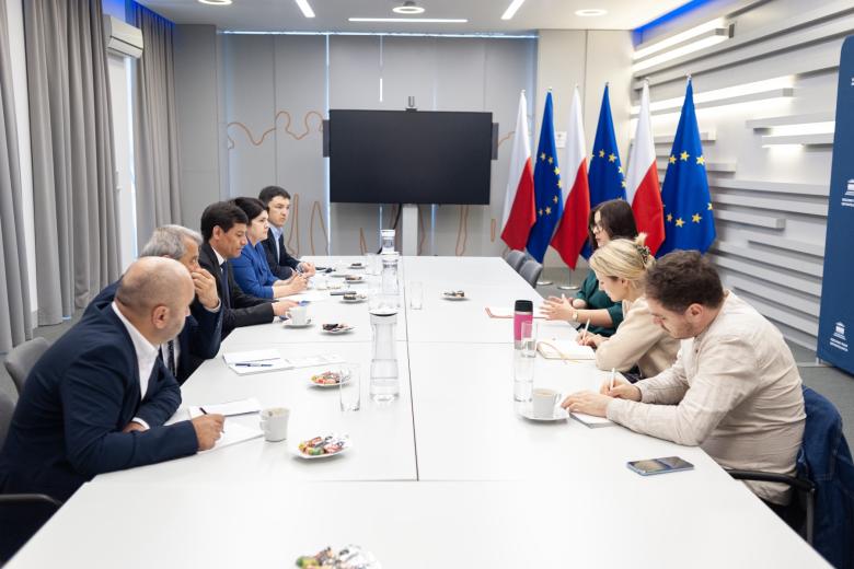 Kilkanaście osób siedzi po dwóch stronach dużego prostokątnego stołu i rozmawia, w tle flagi Polski i UE