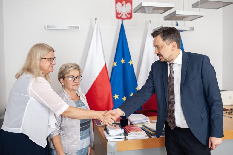 Dwie osoby witają się ściskając dłonie na tle flag Polski i UE
