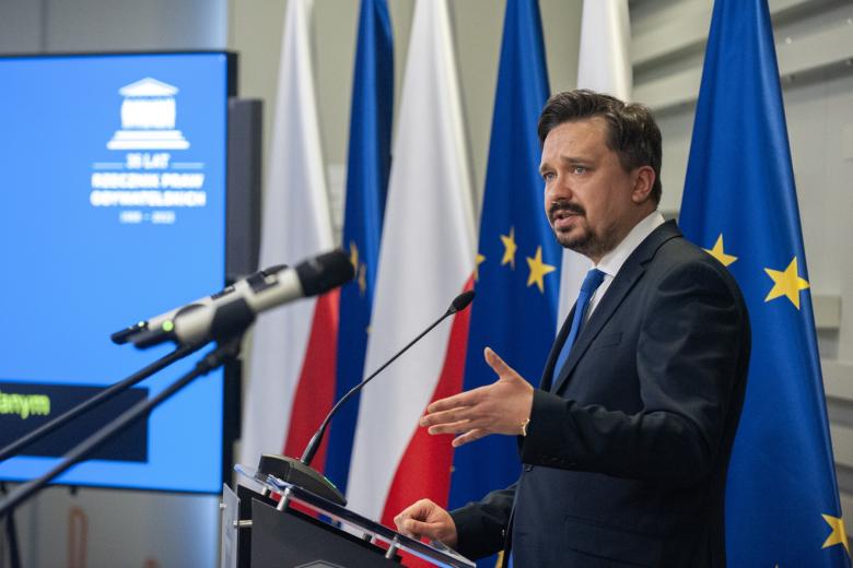 RPO Marcin Wiącek przemawia na stojąco do mikrofonu, w tle flagi Polski i UE