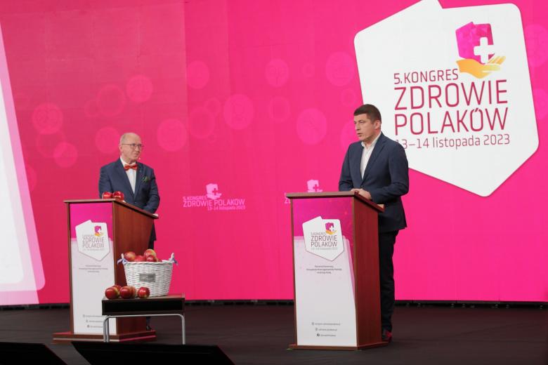 Dwie osoby stoją na scenie za mównicami, jedna przemawia. W tle wyświetlacz z konturem Polski i napisem "Kongres Zdrowie Polaków"