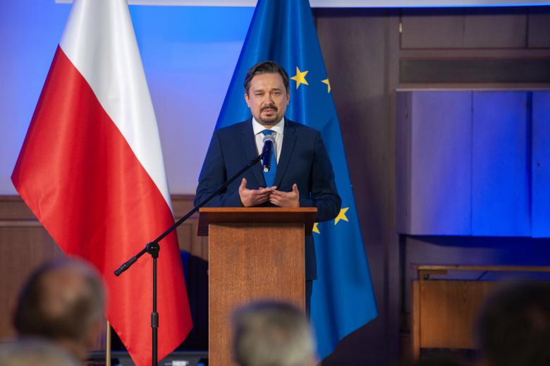 RPO Marcin Wiącek wypowiada się z podium w stronę publiczności. W tle flagi Polski i Unii Europejskiej