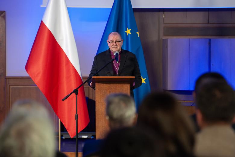 Biskup stojąc przy mównicy wypowiada się w stronę publiczności. W tle flagi Polski i Unii Europejskiej