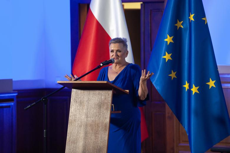 Alicja Szatkowska wypowiada się stojąc przy podium. W tle flagi Polski i Unii Europejskiej