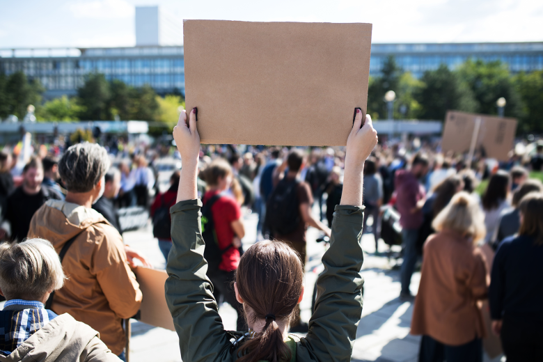 Manifestacja na miejskim placu. Na pierwszym planie stojąca tyłem do kadru kobieta trzymająca tekturowy transparent