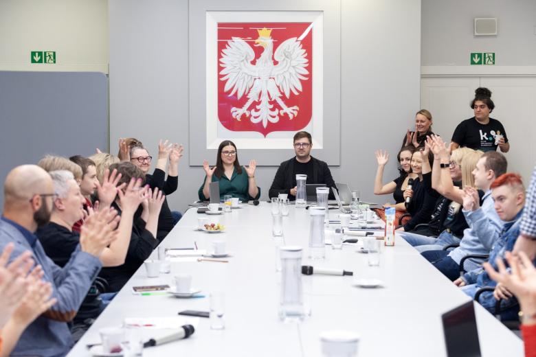 Kilkanaście osób siedzi przy dużym prostokątnym stole konferencyjnym, w tle godło Polski - biały orzeł na czerwonym tle