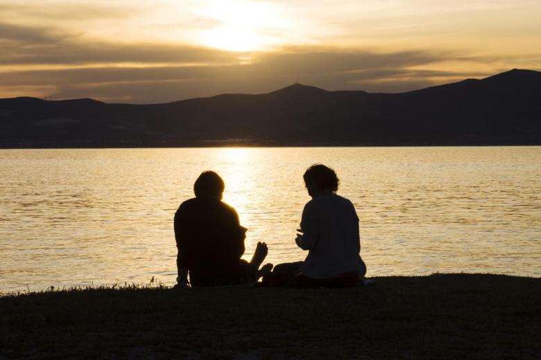 dwie kobiety siedzą tyłem do kamery nad morzem na tle zachodu słońca nad wzgórzami 