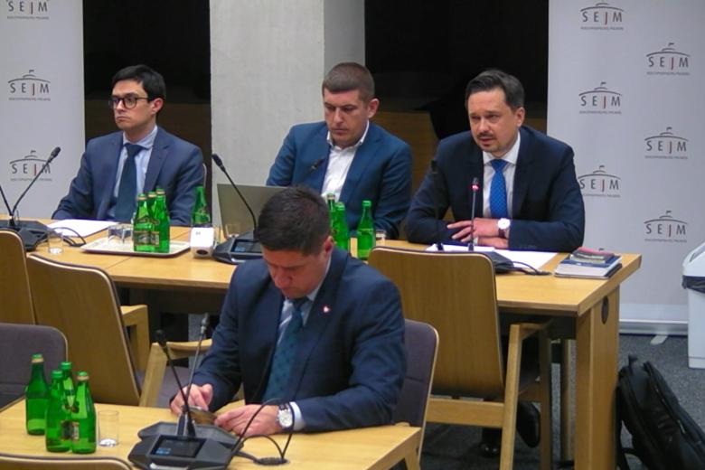 Marcin Wiącek, Wojciech Brzozowski i Michał Szwast siedzą przy stole w sali komisji sejmowej. Marcin Wiącek wypowiada się do mikrofonu.