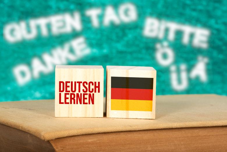Drewniane bloczki z napisem "Deutsch lernen" i flagą Niemiec. W tle szkolna tablica kredowa z zapisanymi niemieckimi słówkami