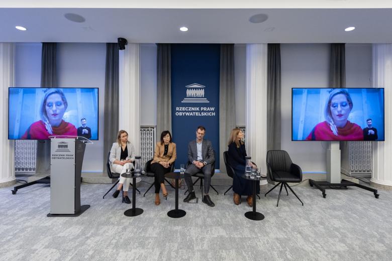 Cztery osoby siedzą na sali konferencyjnej, na telewizorze obok twarz osoby uczestniczącej w spotkaniu za pomocą programu do komunikowania się na odległość