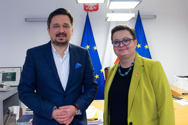 Marcin Wiącek i Katarzyna Lubnauer uśmiechają się pozując do zdjęcia w gabinecie Rzecznika. W tle godło Polski oraz flagi Polski i Unii Europejskiej