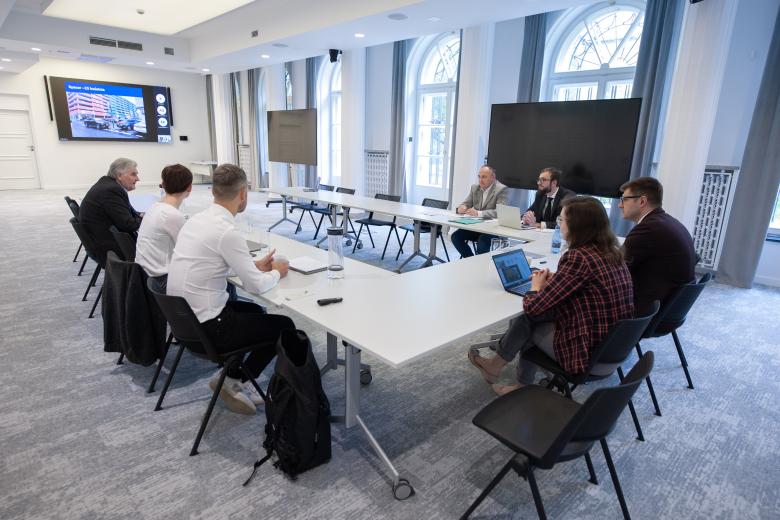 Siedem osób siedzi przy stole konferencyjnym w kształcie podkowy. W szczycie stołu na ekranie wyświetlana jest prezentacja i uczestnicy łączący się za pośrednictwem internetu.