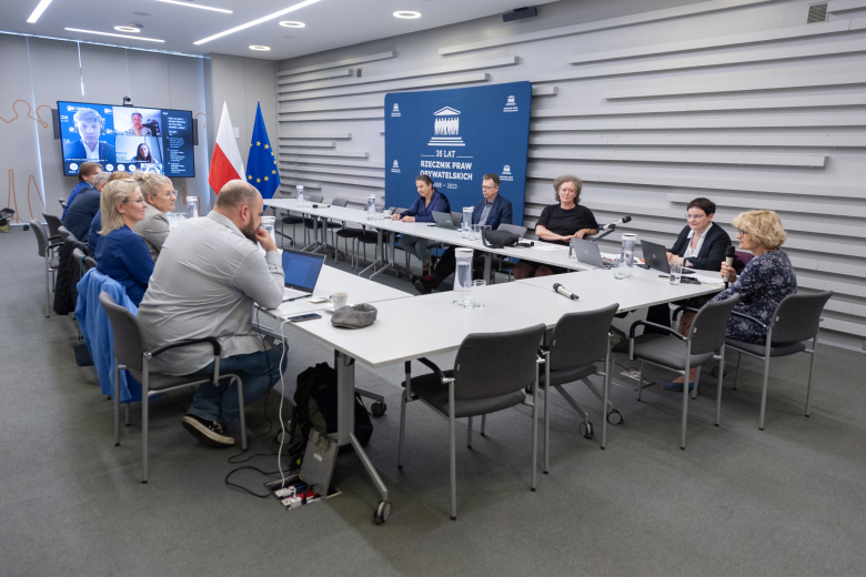 W sali konferencyjnej członkowie komisji siedzą przy ustawionym w kształt podkowy konferencyjnym stole. Na ekranie znajdującym się w szczycie stołu konferencyjnego wyświetleni są również członkowie Komisji biorący udział w spotkaniu za pośrednictwem Internetu. W tle flagi Polski i Unii Europejskiej
