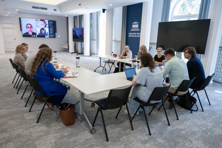 Kilkanaście osób siedzi przy stole konferencyjnym ustawionym w kształt podkowy w sali konferencyjnej, na ścianie wisi duży ekran na którym wyświetlone są osoby uczestniczące w spotkaniu przez internet
