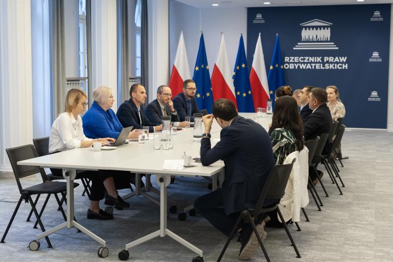 Kilkanaście osób siedzi po dwóch stronach dużego stołu na sali konferencyjnej, w tle granatowa ścianka z białym napisem "Rzecznik Praw Obywatelskich" i flagi Polski i UE