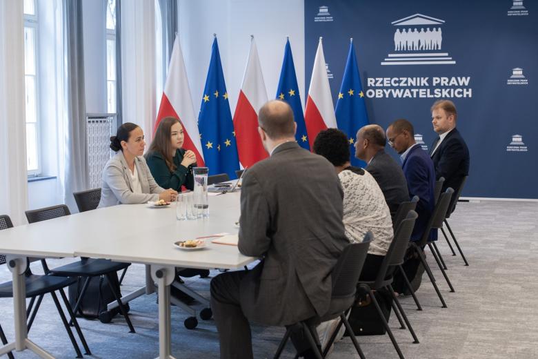 Siedem osób siedzi po obu stronach stołu w sali konferencyjnej i rozmawia. W tle flagi Polski i UE