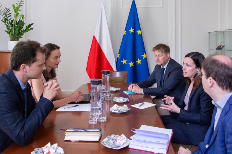Pięć osób rozmawia przy stole w małym pokoju konferencyjnym. W szczycie stołu stoją flagi Polski i Unii Europejskiej