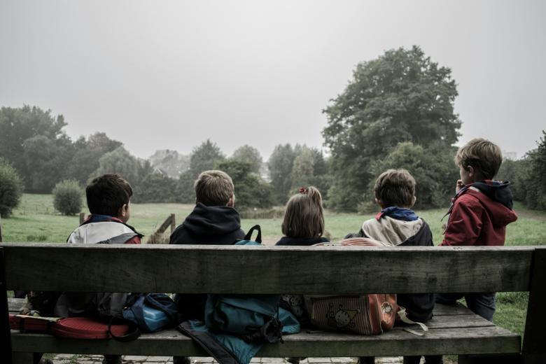 Pięcioro dzieci siedzi na ławce przed łąką i drzewami