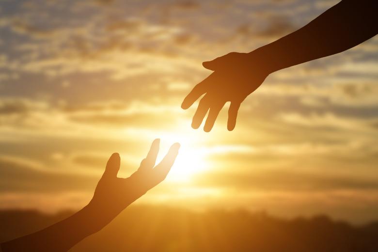 Dwie sięgające do siebie dłonie w geście pomocy na tle zachodzącego słońca