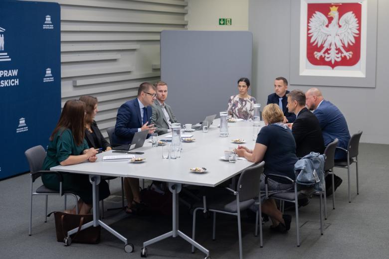 Dziewięć osób siedzi przy dużym prostokątnym stole w sali konferencyjnej i rozmawia, w tle na ścianie duże godło Polski - biały orzeł w koronie na czerwonym polu.