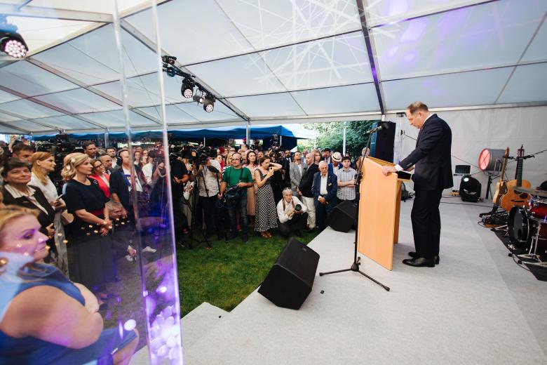 Ambasador USA zwraca się w stronę licznie zgromadzonych gości przyjęcia w dużym namiocie rozstawionym w ogrodzie
