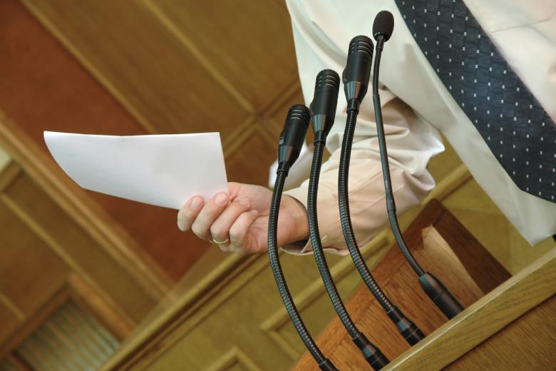 osoba przemawia na stojąco przed nią kilka mikrofonów 