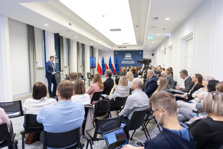 Kilkadziesiąt osób siedzi na sali konferencyjnej i słucha wystąpienia osoby mówiącej za mównicą na środku