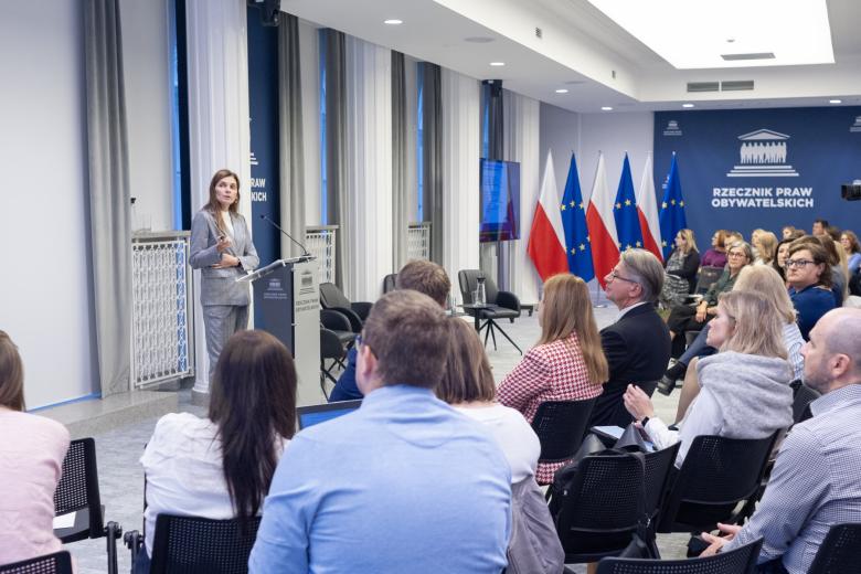 Kilkadziesiąt osób siedzi na sali konferencyjnej i słucha wystąpienia osoby mówiącej za mównicą na środku