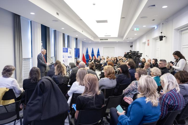 Kilkadziesiąt osób siedzi na krzesłach na sali konferencyjnej i słucha wystąpienia osoby mówiącej za mównicą