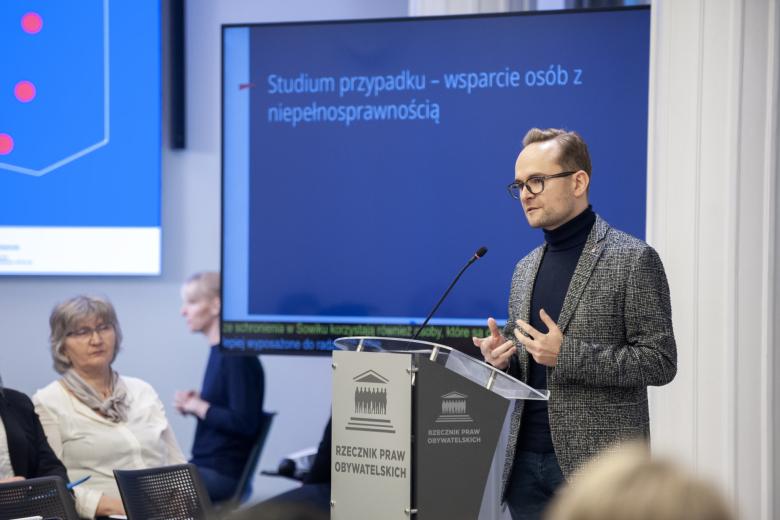 Igor łyko wypowiada się stojąc przy podium. W tle słuchający go uczestnicy konferencji