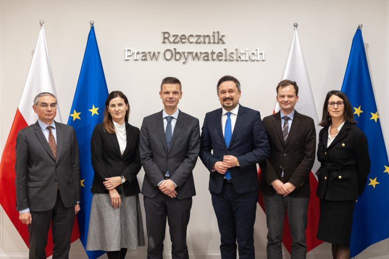 Sześć osób pozuje na stojąco do wspólnego zdjęcia na tle flag Polski i EU oraz napisu "Rzecznik Praw Obywatelskich"