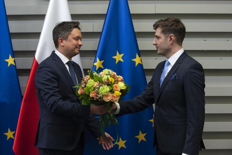 Uśmiechnięty Marcin Wiącek wręcza bukiet kwiatów Valeri'emu Vachevovi na tle flag Polski i Unii Europejskiej