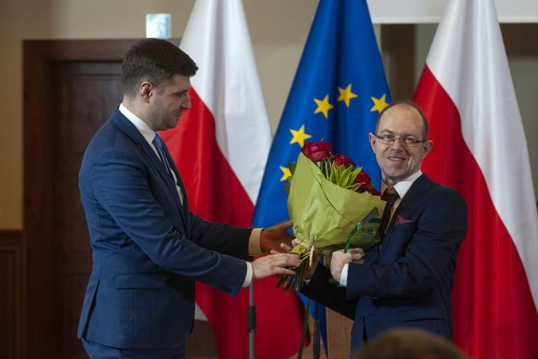 Valeri Vachev wręcza kwiaty uśmiechającemu się Krzysztofowi Kurowskiemu na tle flag Polski i Unii Europejskiej