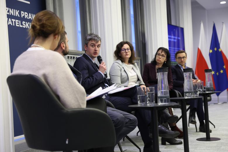 Uczestnik panelu trzyma mikrofon i wypowiada się w stronę siedzących obok niego innych uczestników panelu