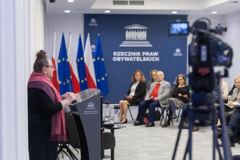 Sala konferencyjna. Barbara Imiołczyk i siedzący uczestnicy debaty