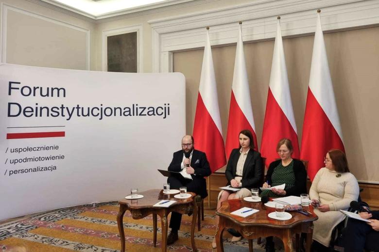 Cztery osoby siedzą na krzesłach przy małych stolikach, jedna z nich mówi do mikrofonu