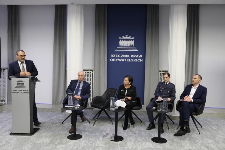 Osoba wypowiada się, stojąc przy mównicy. Obok niej siedzi czterech panelistów.