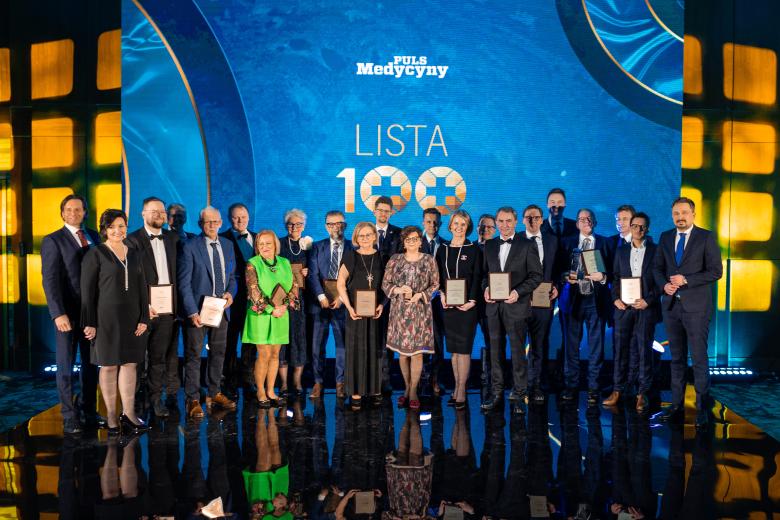 Grupa osób elegancko ubranych pozuje na scenie do zdjęcia. W tle widać ekran z napisem "Puls Medycyny. Lista 100"