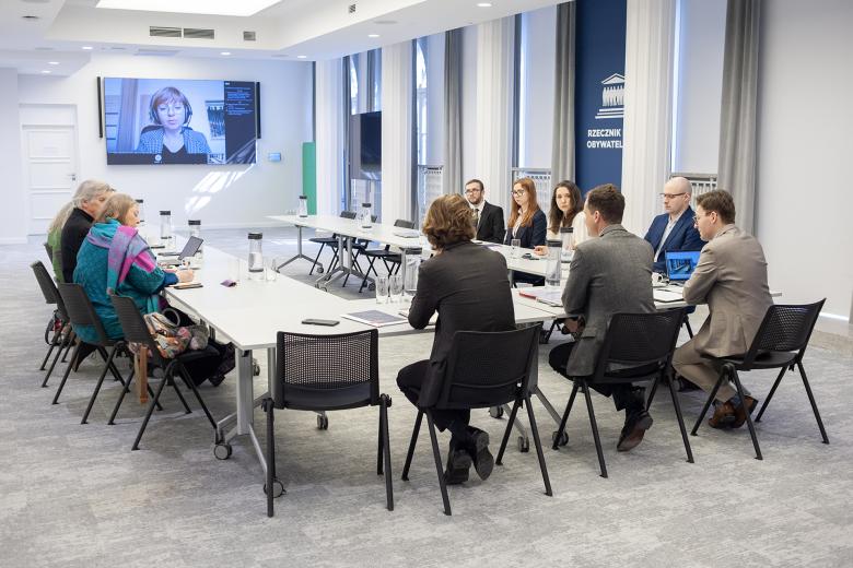 Kilkanaście osób siedzi przy stołach ustawionych w podkowę, na dużym telewizorze na ścianie widoczni uczestnicy posiedzenia łączący się zdalnie za pomocą programu do komunikowania się na odległość