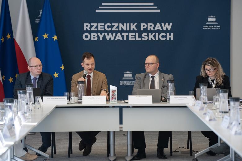 Cztery osoby siedzą w szczycie stołu konferencyjnego. Jedna z nich wypowiada się do trzymanego w dłoniach mikrofonu