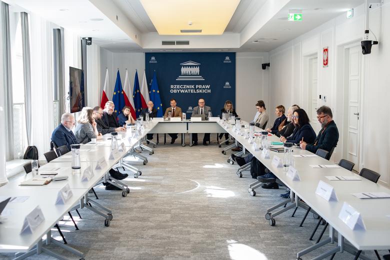 Kilkanaście osób siedzi przy stołach konferencyjnych ustawionych w kształt podkowy.