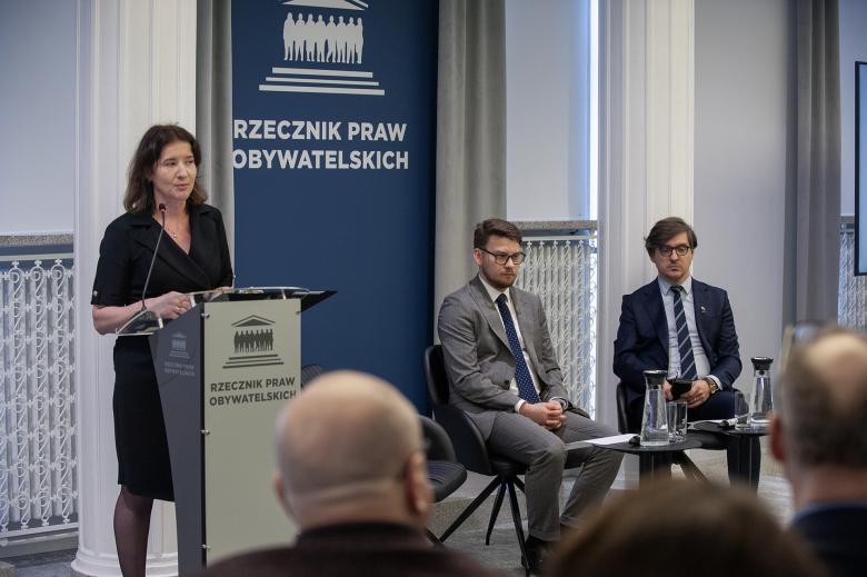 Marzena Andrzejewska wypowiada się do mikrofonu stojąc przy mównicy. Obok niej na krzesłach siedzą inni uczestnicy panelu. Na pierwszym planie widać sylwetki osób z widowni