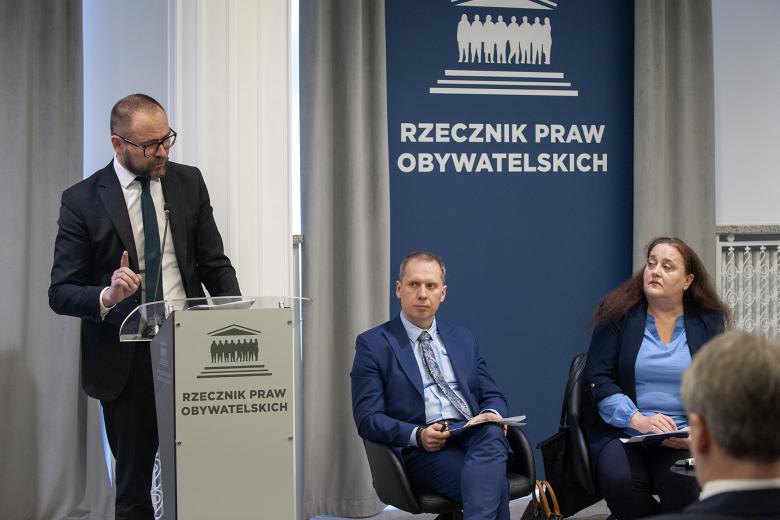 Jarosław Zagrodnik wypowiada się do mikrofonu stojąc przy mównicy i gestykulując. Obok niego siedzą dwie inne osoby uczestniczące w panelu