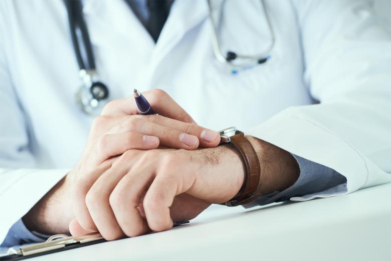 Zbliżenie na położone na blacie biurka dłonie mężczyzny w białym fartuchu i z przewieszonym przez szyję stetoskopem. Mężczyzna sprawdza na zegarku godzinę.