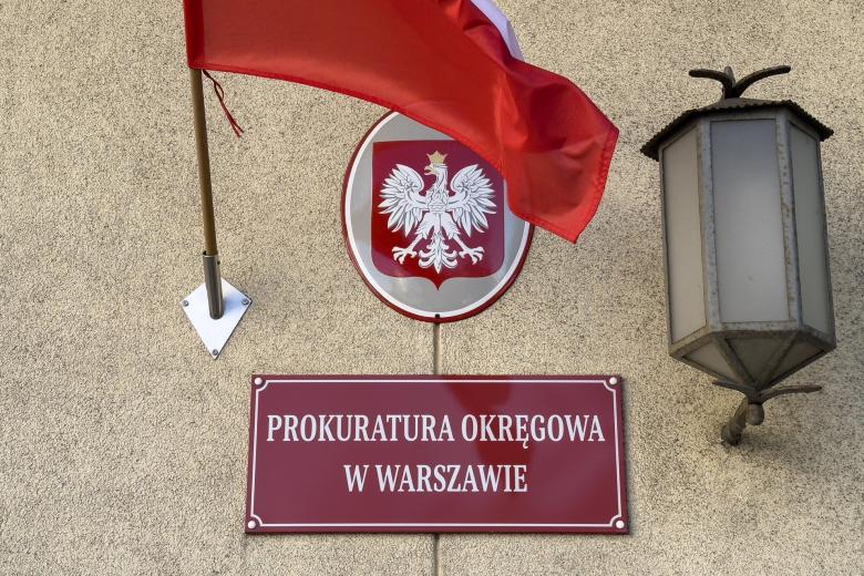 Czerwona tabliczka z białym napisem "PROKURATURA OKREGOWA W WARSZAWIE" wisząca na ścianie budynku, obok biało-czerwona flaga