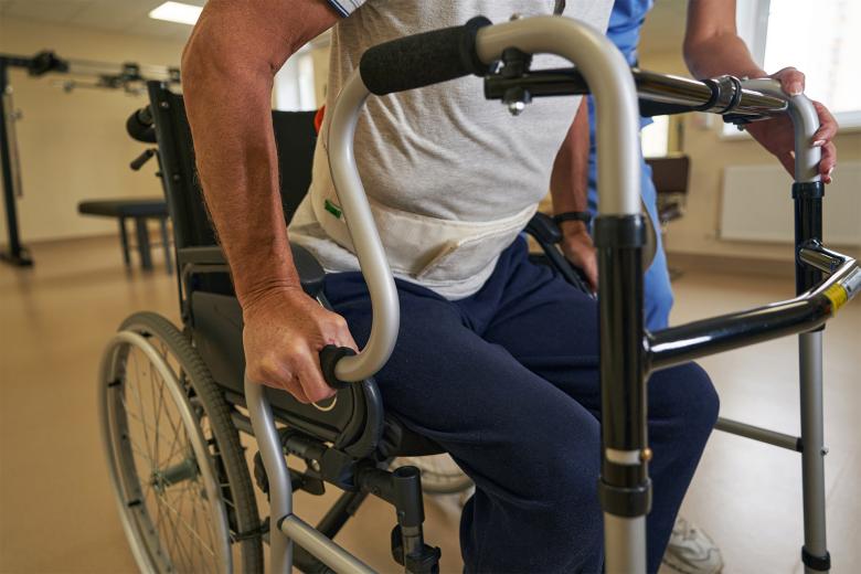 Mężczyzna siedzący na wózku podczas ćwiczeń rehabilitacyjnych. Asystuje mu kobieta w stroju medycznym.