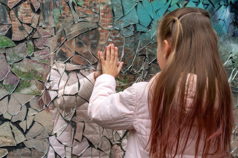 Stojąca tyłem do kadru dziewczynka trzyma dłoń na stojącym przed nią murze wyłożonym odłamkami lustra