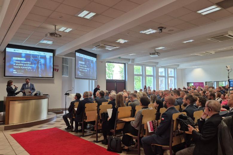 Zapełniona siedzącymi ludźmi aula. Przed miejscami siedzącymi znajduje się audytorium, przy którym stoją dwie osoby. Nad audytorium jest ekran projektora wyświetlający tytuł wydarzenia "Prawa człowieka w służbie publicznej. Teoria a praktyka"