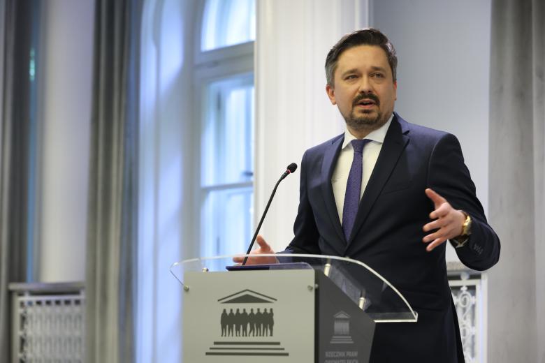 RPO Marcin Wiącek wypowiada się i gestykuluje, stojąc przy mównicy