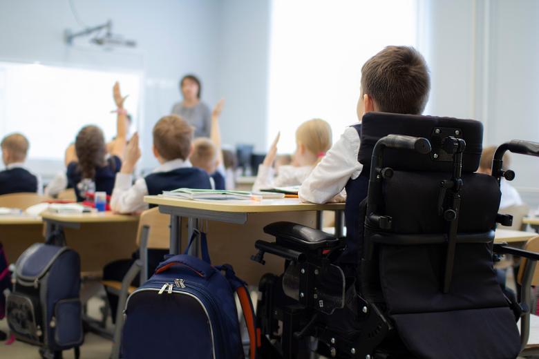 Dziecko siedzące na wózku przy ławce w sali szkolnej pośród innych dzieci, które słuchają nauczyciela
