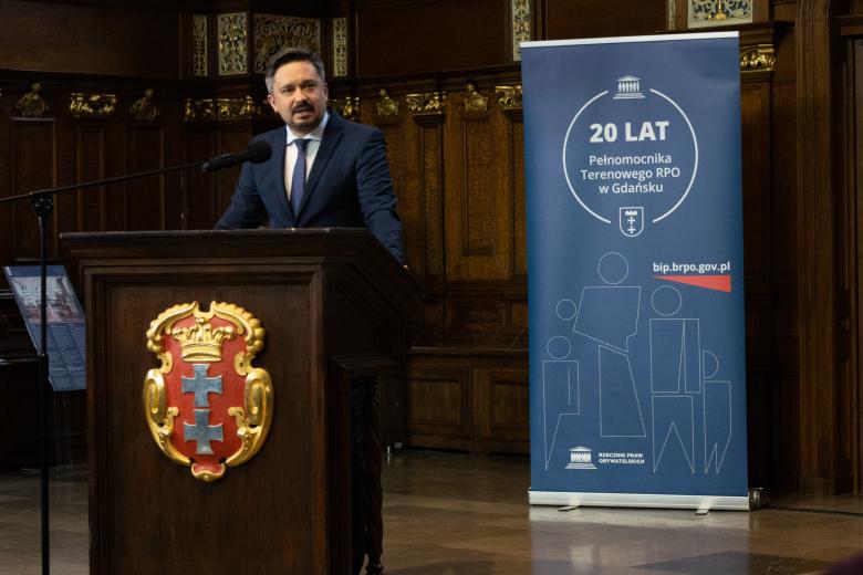 Osoba przemawia za zdobioną, drewnianą mównicą z herbem gdańska - na czerwonym polu złota korona otwarta i dwa równoramienne srebrne krzyże w słup 